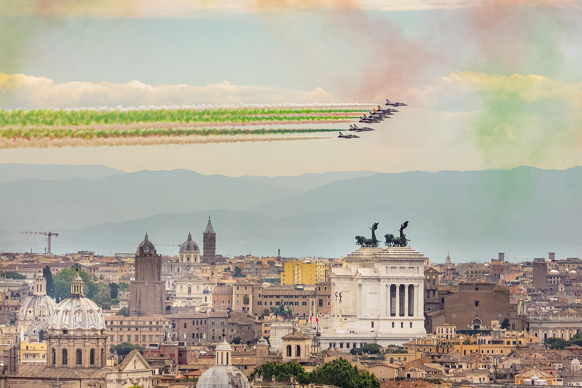 The Frecce Tricolori in nine-jet formation trailing green, white and red smoke over the Altare della Patria in Rome on 2 June 2021. The cityscape of Rome, including the Vittoriano monument with its bronze chariot, is visible below the formation.