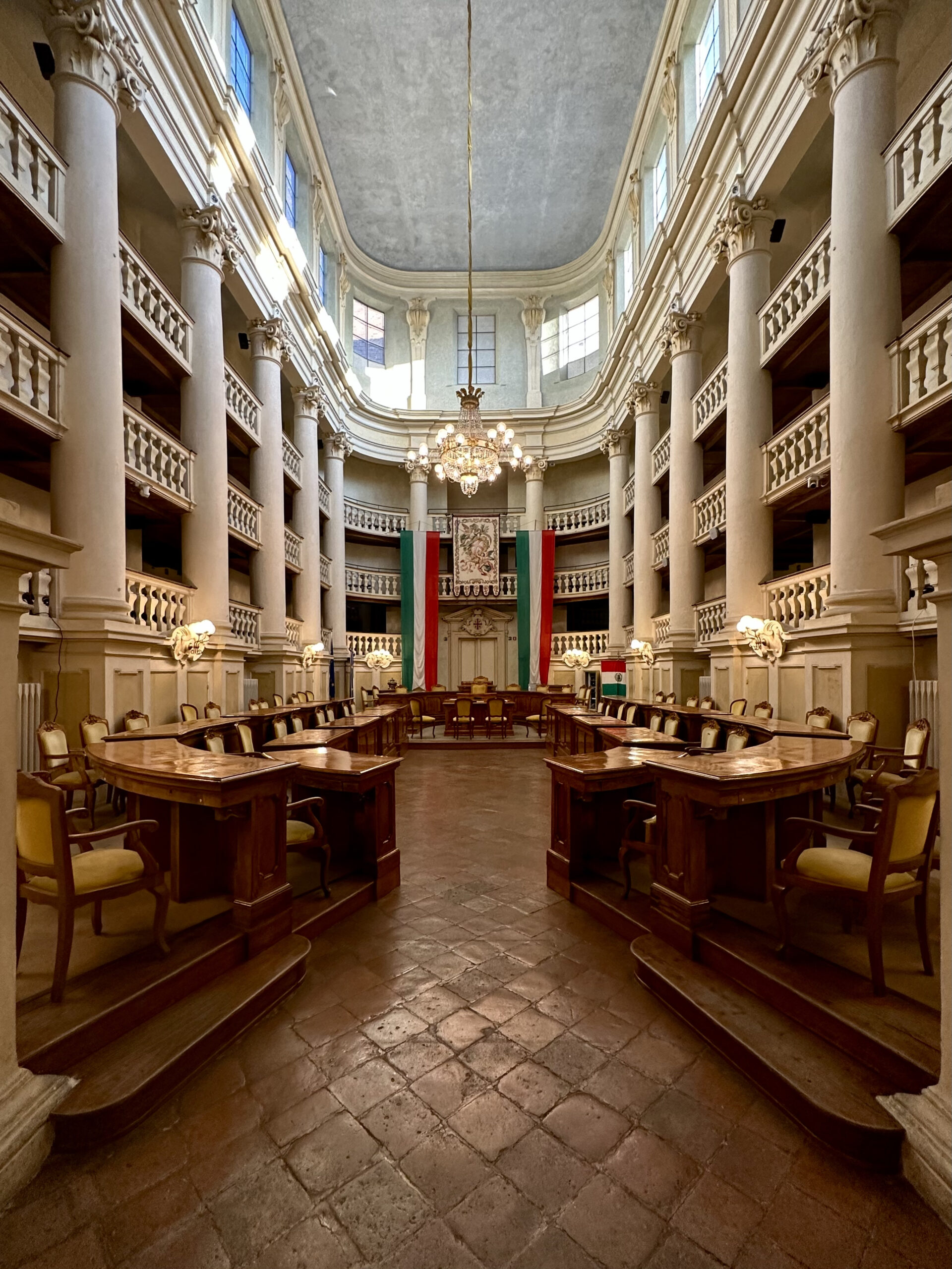 Interior of the Sala del Tricolore in Reggio Emilia, the neoclassical chamber where the Cispadane Congress voted to adopt the green-white-red flag on 7 January 1797. Empty council benches arranged in a horseshoe, with the historical tricolore visible at the rear.